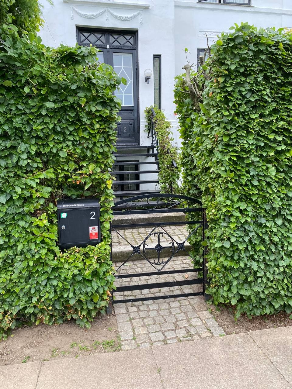 Indgang med smedejernslåge, brostensbelagt sti og klippede hække ved hvid villa