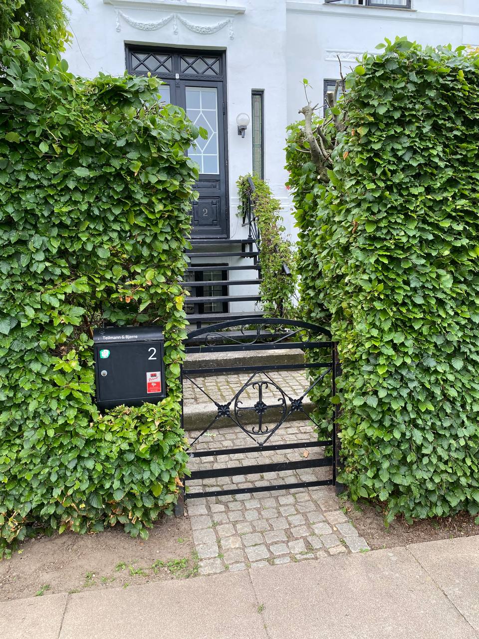 Indgang med smedejernslåge, brostensbelagt sti og klippede hække ved hvid villa
